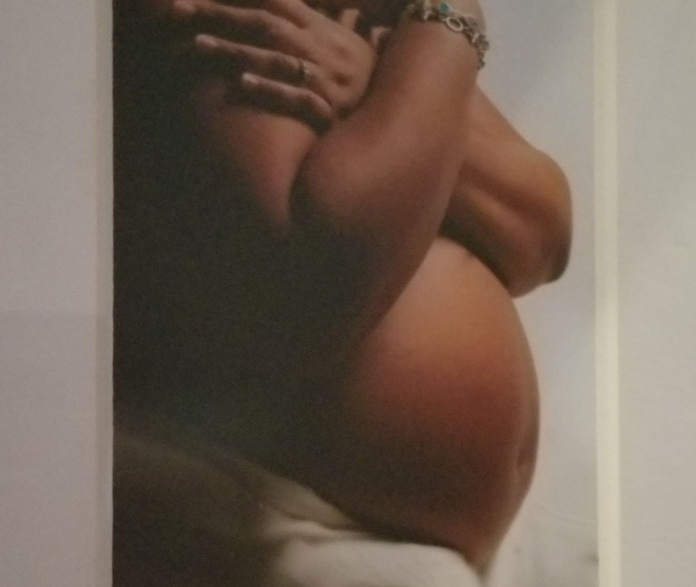 Brown skinned, bare pregnant belly with mother's hands crossed over her breasts and a white background