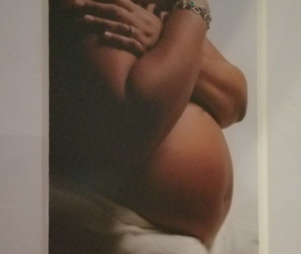 Brown skinned, bare pregnant belly with mother's hands crossed over her breasts and a white background